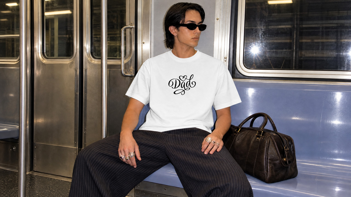 A stylish person wearing sunglasses and a white T-shirt with a black “Dad” script design sits inside a subway car beside a black handbag.