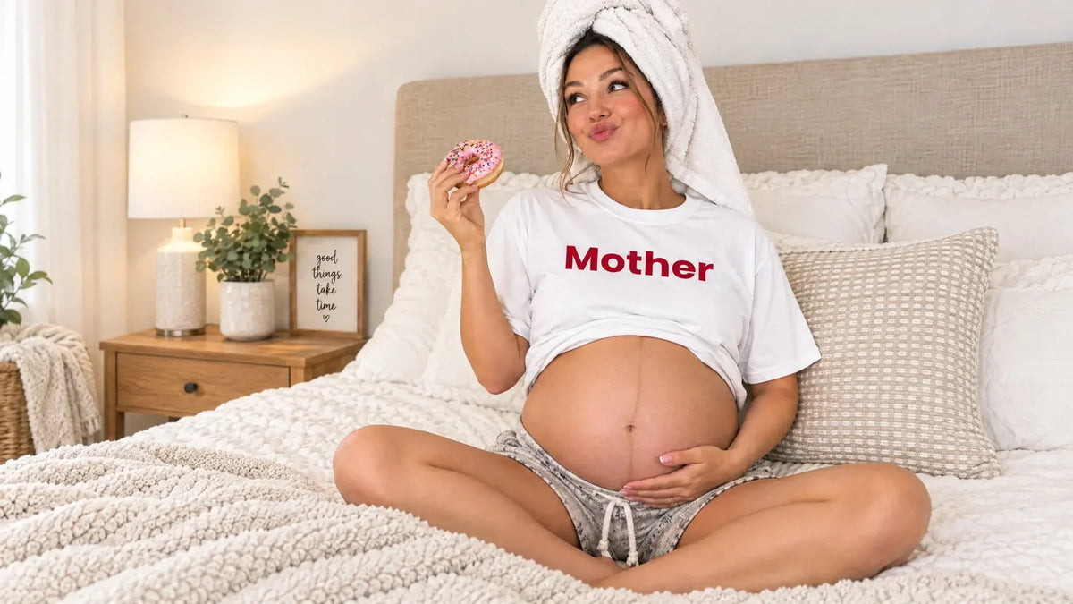 A pregnant woman wearing a white “Mother” T-shirt and a towel wrapped around her hair sits on a cozy bed holding a pink-frosted donut