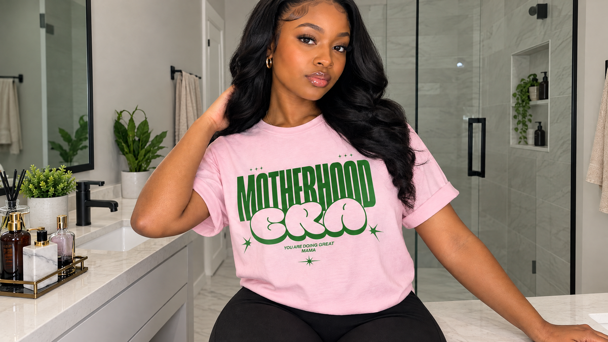 A woman with long black hair wearing a pink “MOTHERHOOD ERA” T-shirt stands beside a modern bathroom vanity, with the shirt message “YOU ARE DOING GREAT MAMA” visible below the main design.
