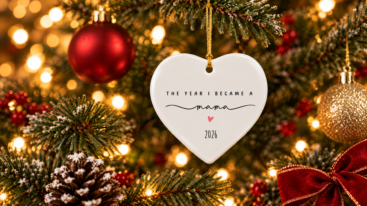 A white heart-shaped Christmas ornament hanging on a decorated tree, featuring the message “THE YEAR I BECAME A mama,” a pink heart, and the year “2026,” surrounded by glowing lights, evergreen branches, red ornaments, and a festive red bow.